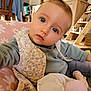 baby, child, face, blue_eyes, vest, green_shirt, indoor, home, toy, plush, furniture, wooden_chair, shelf, decor, photography, portrait, curious, candid, seated, closeup