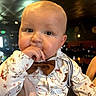 Silas is registered to the contest to win money with this photo: baby, child, face, hand, finger, bow_tie, shirt, floral_pattern, suspenders, indoor, restaurant, lighting, portrait, cute, person, young_child, expression, clothing, holding, warm_lighting