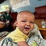 baby, smiling, yellow_shirt, blanket, indoor, chubby_cheeks, happy, face, person, child, toddler, soft_texture, portrait, cute, lying_down, cozy, warm_light, closeup, infant, expression