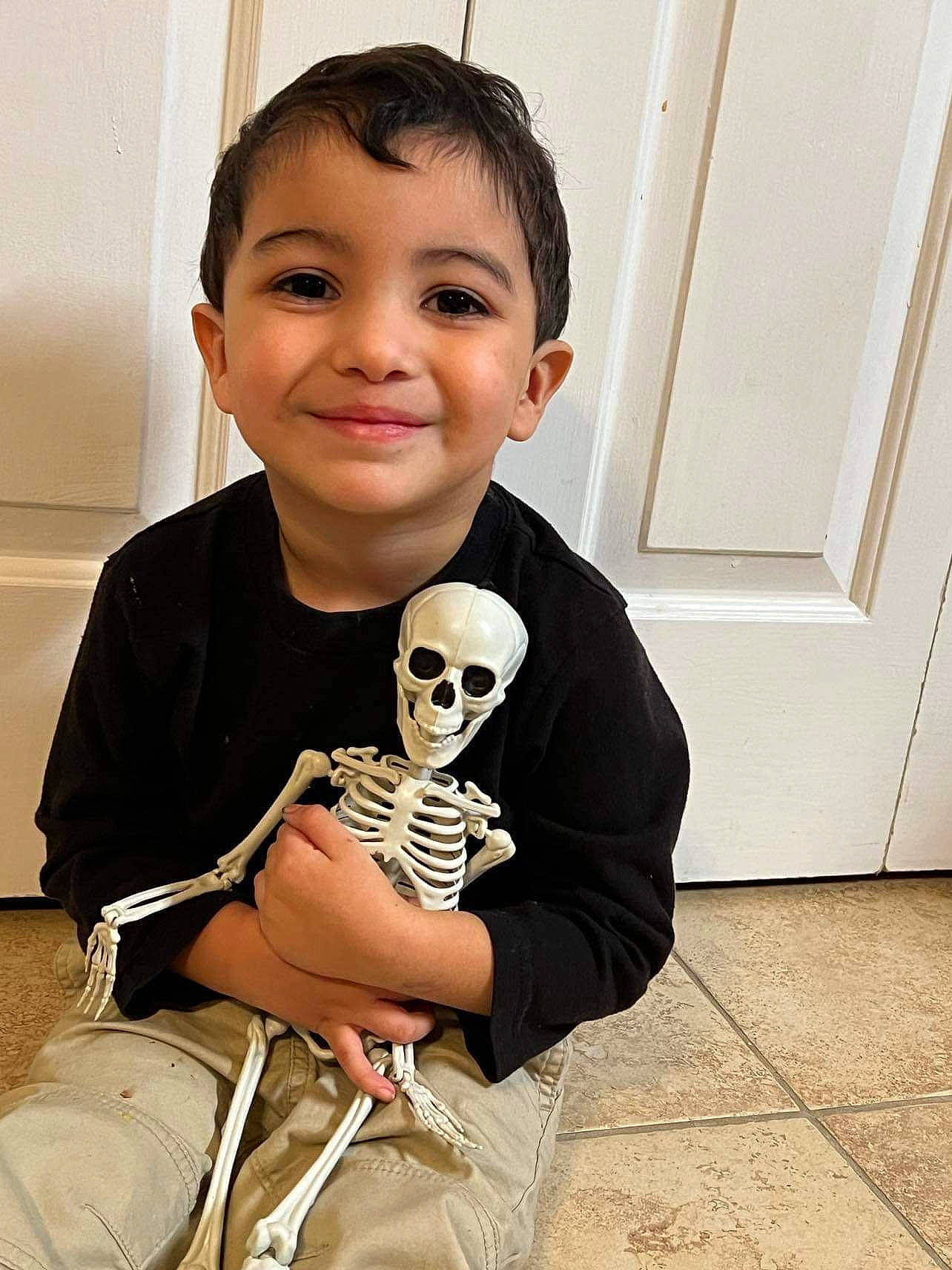 Lance is registered to the contest to win money with this photo: baby_toddler_clothing, bone, child, flooring, fun, happy, head, human_body, joy, pattern, person, personal_protective_equipment, room, sitting, skeleton, skull, sleeve, smile, sneakers, t_shirt
