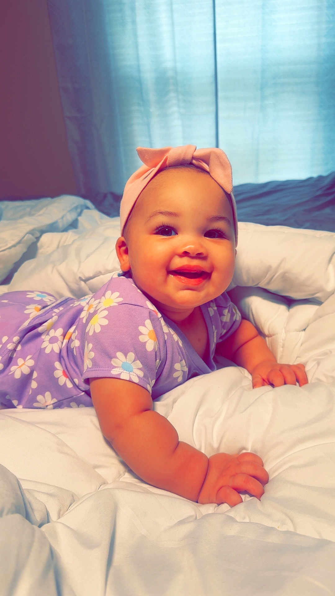 Aurorah is registered to the contest to win money with this photo: baby, bedroom, child, chubby_arms, closeup, comforter, curtains, cute, daisy_pattern, happy, headband, indoor, infant, laying, person, portrait, purple_dress, smiling, soft_light, white_bedding