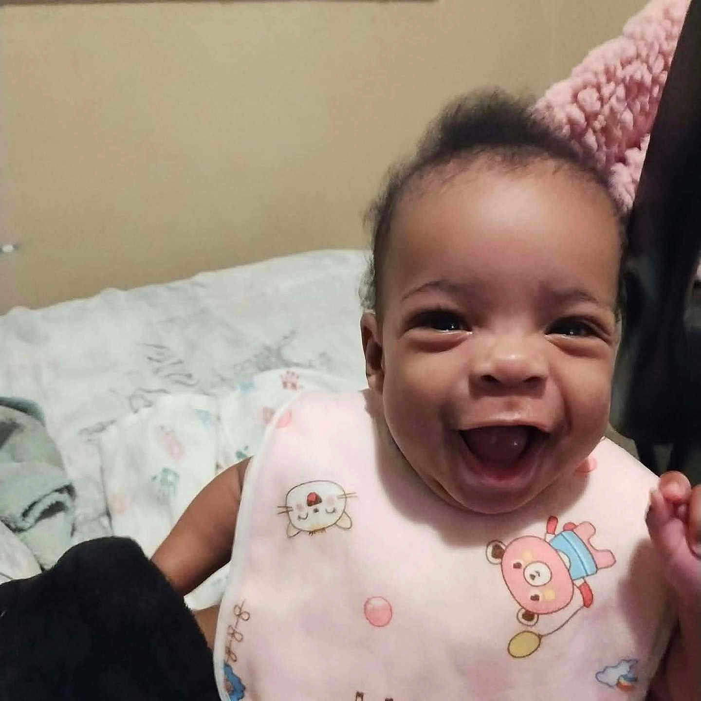 Iyana is registered to the contest to win money with this photo: baby, child, infant, smiling, laughing, portrait, indoors, bedroom, bed, bedding, pink_clothing, animal_print, onesie, happy, face, head, dark_hair, sitting, candid, family_photo