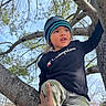 beanie, branches, camouflage_pants, casual_clothing, champion, child, climbing, curious, daylight, hand, nature, outdoor, person, portrait, sky, support, sweatshirt, tattoo, tree, young