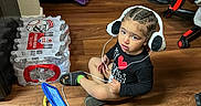 Keshaun is registered to the contest to win money with this photo: black_clothing, blanket, bottled_water, braided_hair, child, curious_expression, flooring, furniture, headphones, indoor, living_room, packaging, person, playtime, sitting, sock, tablet, technology, toddler, wooden_floor