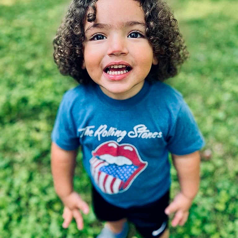 Keshaun is registered to the contest to win money with this photo: blue_shirt, casual_clothing, child, curly_hair, cute, daylight, fun, grass, happy, outdoor, person, portrait, rolling_stones, shoes, shorts, smiling, standing, summer, toddler, young_child