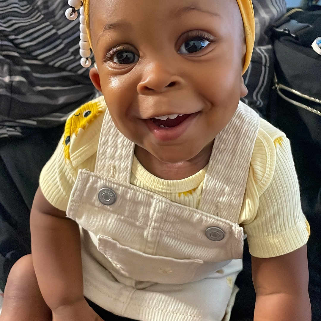 Autumn J is registered to the contest to win money with this photo: baby, beads, child, clothing, cushion, cute, eyes, face, floral_pattern, hands, happy, indoors, legs, overalls, portrait, sitting, smiling, toddler, yellow_headband, yellow_shirt