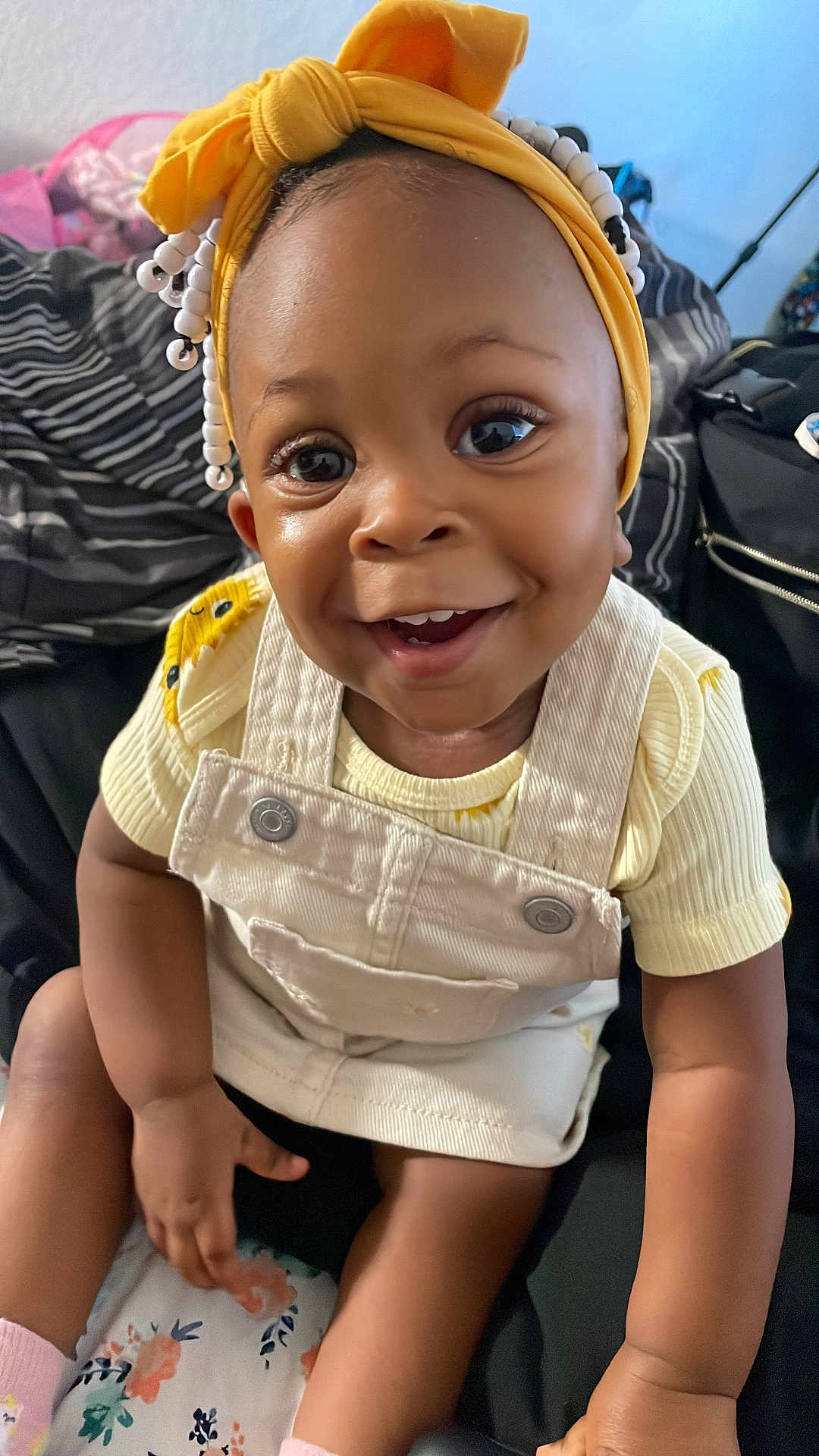Autumn J is registered to the contest to win money with this photo: toddler, child, smiling, yellow_headband, beads, overalls, yellow_shirt, sitting, indoors, cushion, floral_pattern, happy, cute, face, eyes, hands, legs, clothing, portrait, baby