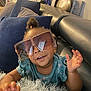 toddler, child, sunglasses, blue_shirt, couch, pillows, rug, home_interior, smiling, happy, person, indoor, furniture, hair_tied, playful, leather_couch, hand, face, living_room, cozy