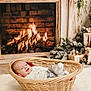 baby, basket, fireplace, fire, cozy, holiday, decorations, gift, wrapped_gift, plush_rug, smiling, infant, warm, indoors, wood, wicker, blanket, clothing, socks, festive