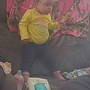 Aliyah is registered to the contest to win money with this photo: child, toddler, standing, yellow_shirt, pink_socks, couch, blanket, living_room, window, blinds, water_bottle, wipes, container, household, indoor, portrait, face, clothing, furniture, home
