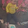 child, toddler, standing, yellow_shirt, pink_socks, couch, blanket, living_room, window, blinds, water_bottle, wipes, container, household, indoor, portrait, face, clothing, furniture, home