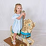 background, blonde_hair, blue_dress, carousel_horse, child, childhood, cute, floor, fun, girl, happy, indoor, play, pole, portrait, smile, toy, vintage, white_sandals, wooden_base