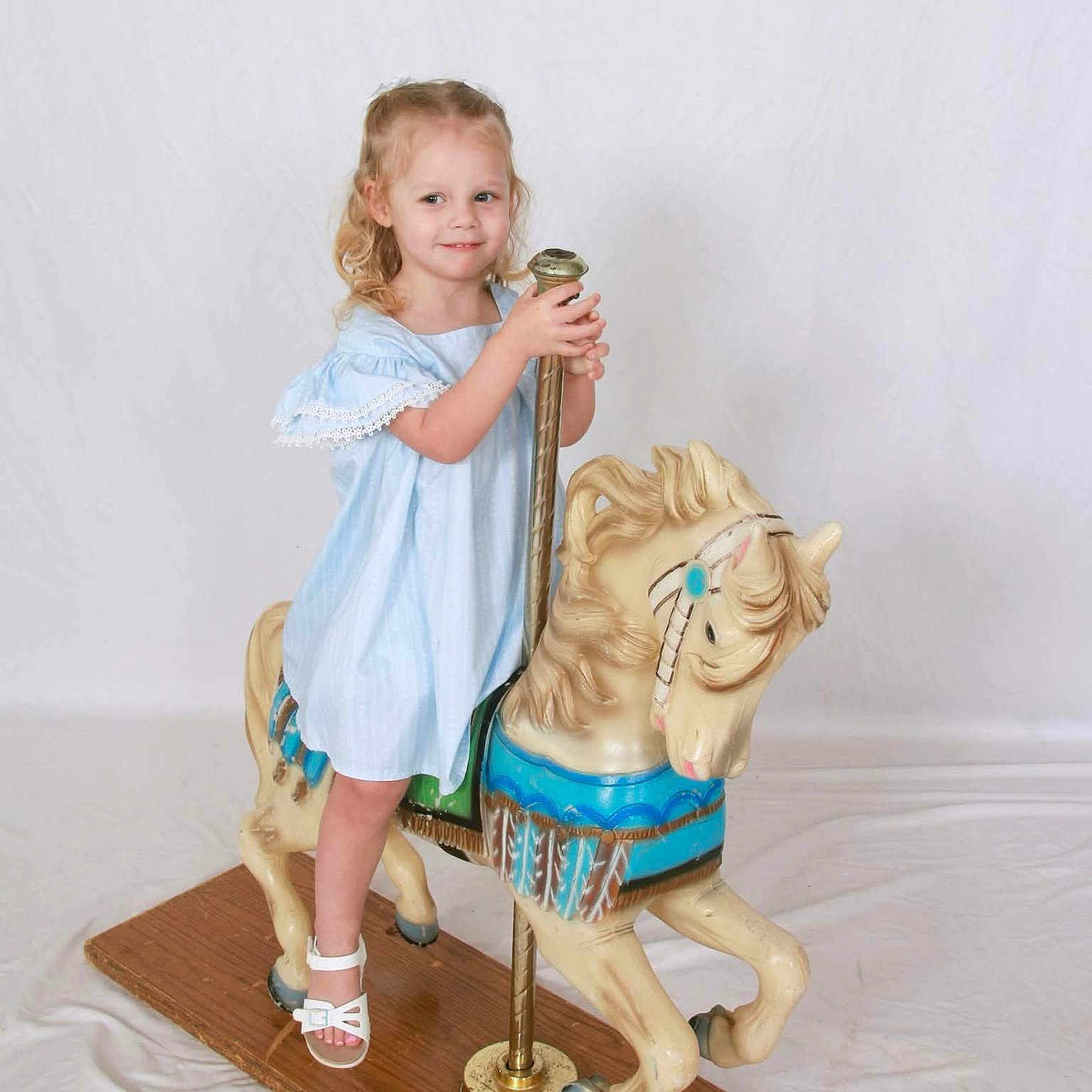 Presleigh joined the competition — help win amazing prizes! background, blonde_hair, blue_dress, carousel_horse, child, childhood, cute, floor, fun, girl, happy, indoor, play, pole, portrait, smile, toy, vintage, white_sandals, wooden_base