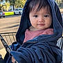 toddler, child, shopping_cart, hooded_jacket, blue_clothing, outdoor, daylight, trees, face, eyes, curious, street, parked_cars, clothing, person, portrait, cute, infant, baby, smiling