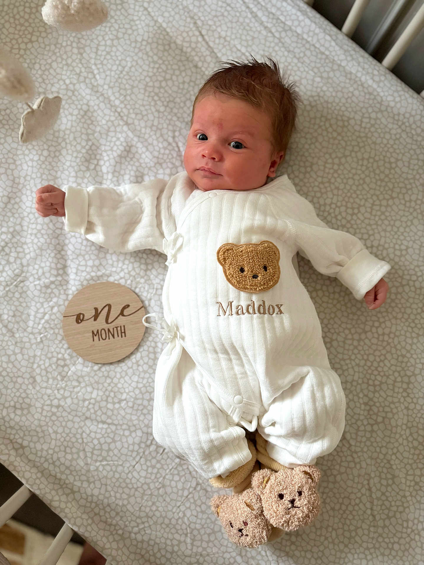 Maddox is registered to the contest to win money with this photo: baby, infant, newborn, onesie, teddy_bear, slippers, crib, blanket, wooden_sign, milestone, cute, white_clothing, patterned_bed, lying_down, child, portrait, indoor, soft_lighting, sleepwear, baby_boy