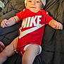 baby, infant, smiling, red_clothing, nike_logo, baby_bottle, pacifier, blanket, lying_down, hat, sock, cute, happy, indoor, child, person, portrait, soft_lighting, comfort, playful