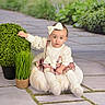 baby, bow, bush, child, cushion, dress, floor_tiles, girl, grass, hand, headband, infant, plush, portrait, pose, potted_plant, sitting, socks, studio_backdrop, surprised_expression
