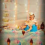 baby, child, crown, blue_seat, pink_dress, tablecloth, ice_cream_cone, ice_cream_tub, hersheys_syrup, string_lights, party, birthday, decorations, cupcakes, bowl, food, colorful, indoor, celebration, cute