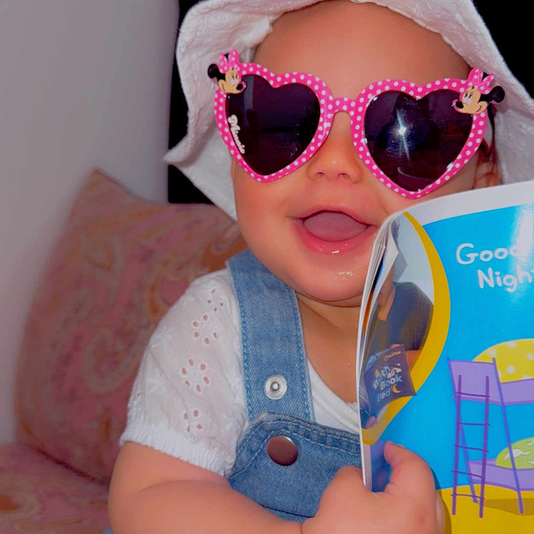 Evellyn joined the competition — help win amazing prizes! baby, book, child, clothing, couch, cute, denim_overalls, happy, hat, heart_shaped, indoor, minnie_mouse, person, pink, portrait, reading, seated, smiling, sunglasses, white_hat