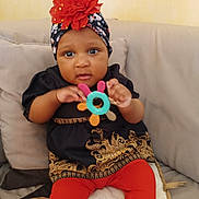 Assiah participe au concours pour gagner de l'argent avec cette photo : baby, black_clothing, child, clothing, colorful_toy, couch, curious, cute, flower, furniture, gold_pattern, headwrap, indoor, infant, person, portrait, red_leggings, sitting, teething_toy, young_child