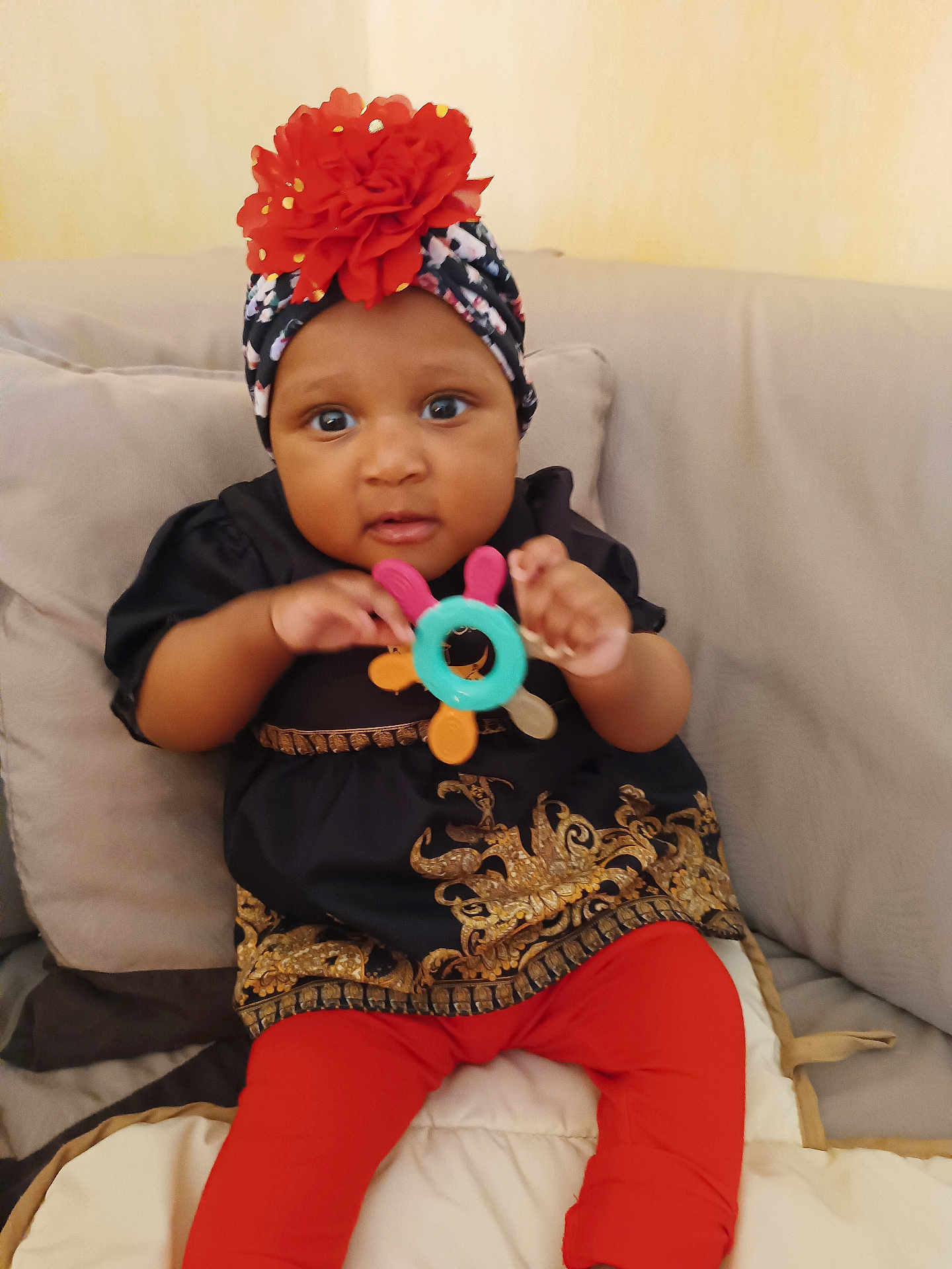 Assiah participe au concours pour gagner de l'argent avec cette photo : baby, black_clothing, child, clothing, colorful_toy, couch, curious, cute, flower, furniture, gold_pattern, headwrap, indoor, infant, person, portrait, red_leggings, sitting, teething_toy, young_child