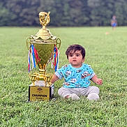 Aaryansh is registered to the contest to win money with this photo: baby, child, toddler, trophy, champion, outdoor, grass, field, sports, football, portrait, sitting, curious, face, hair, blue_shirt, panda_pattern, gray_pants, white_shoes, ribbons