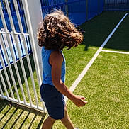 Zwak joined the competition — help win amazing prizes! child, curly_hair, walking, sports_field, green_grass, white_fence, blue_tank_top, shorts, sneakers, sunlight, shadow, outdoor, daylight, playground, athletic, young, person, casual_clothing, side_view, movement