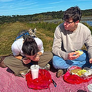 Oak is registered to the contest to win money with this photo: cat, people, picnic, outdoors, lake, water, grass, hillside, harness, leash, checkered_blanket, sandwich, food, drinks, plastic_bags, sunglasses, glasses, sitting, sunny, landscape