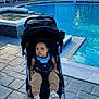toddler, stroller, pool, outdoor, child, building, pavement, sneakers, casual_clothing, daylight, residential_area, curious, sitting, baby, handrail, plant, window, blue, sunlight, relaxation