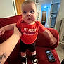 baby, child, red_couch, red_shirt, black_shorts, sneakers, hand, tattoo, smartphone, living_room, door, kitchen, floor, person, standing, indoor, furniture, motion_blur, support, home