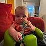 baby, child, toy, green_seat, indoor, living_room, red_couch, door, hallway, kitchen, person, infant, clothing, furniture, holding, seated, curious, face, expression, home