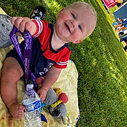 Noel joined the competition — help win amazing prizes! baby, child, grass, blanket, water_bottle, toy, plush_toy, smiling, outdoor, sunny, happy, lanyard, red_clothing, blue_clothing, event, people, park, daylight, cute, sitting