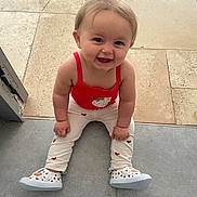 Angèle a rejoint le concours — aidez-le/la à gagner de superbes lots ! baby, child, smiling, sitting, floor, red_top, white_pants, heart_pattern, strawberry_design, shoes, indoor, happy, cute, person, portrait, toddler, young_child, face, human, casual_clothing