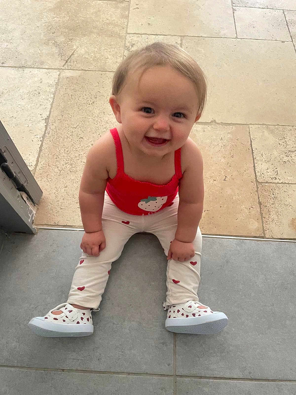 Angèle a rejoint le concours — aidez-le/la à gagner de superbes lots ! baby, child, smiling, sitting, floor, red_top, white_pants, heart_pattern, strawberry_design, shoes, indoor, happy, cute, person, portrait, toddler, young_child, face, human, casual_clothing