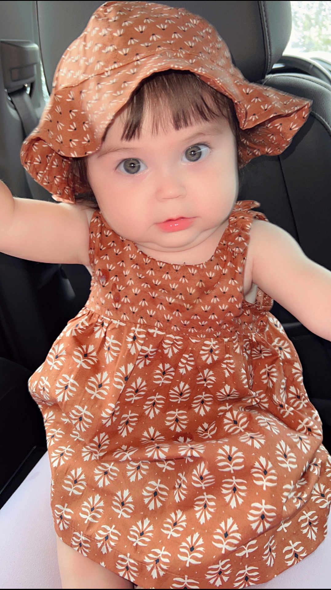 Giselle joined the competition — help win amazing prizes! baby, child, car_seat, hat, dress, patterned_clothing, cute, infant, face, eyes, chubby_cheeks, sitting, indoors, portrait, person, headwear, clothing, seat, young_child, adorable