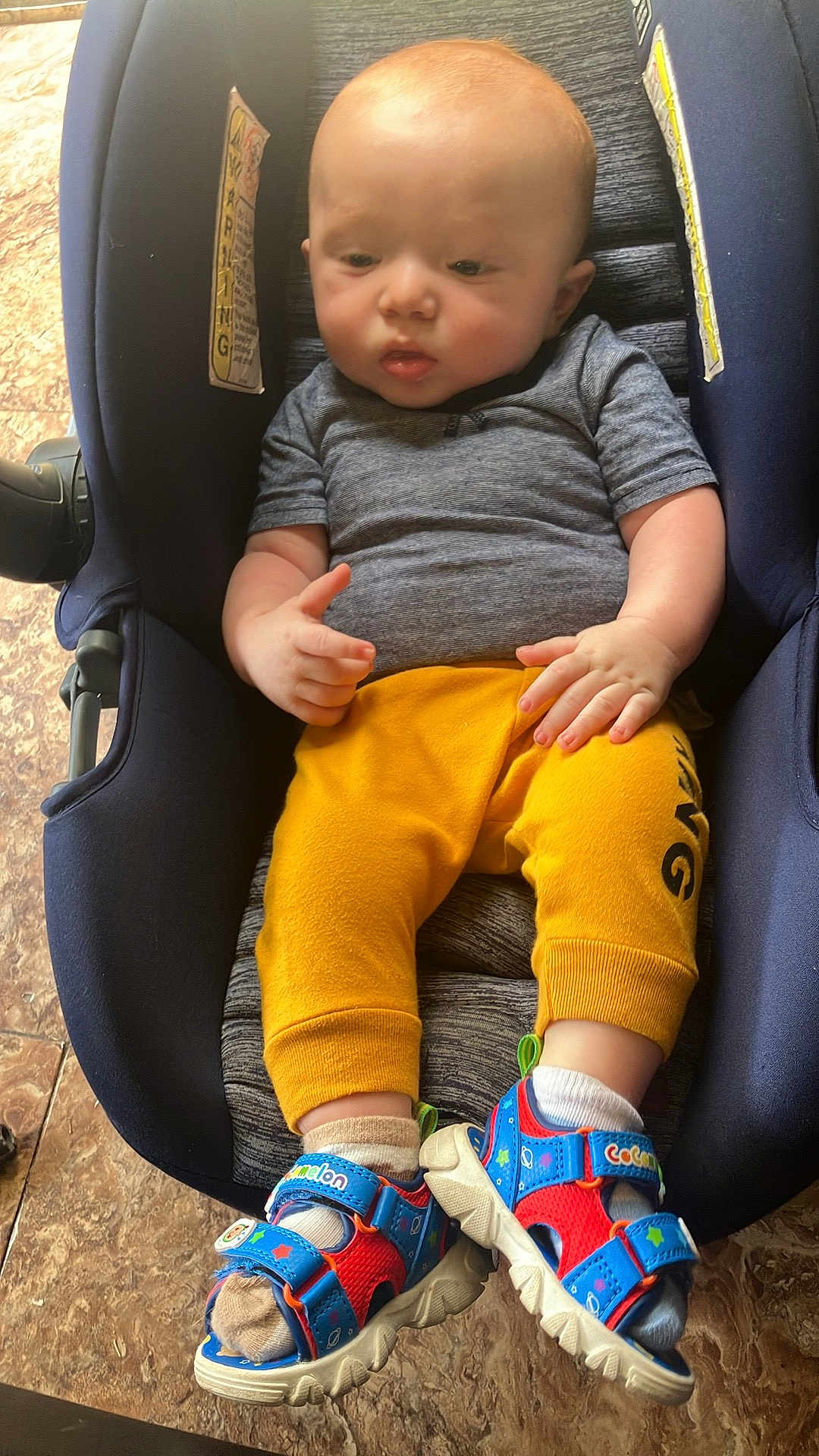 Jaxson is registered to the contest to win money with this photo: baby, infant, car_seat, child, toddler, yellow_pants, grey_shirt, colorful_sandals, socks, floor, tile_floor, indoor, footwear, seated, cute, small_hands, young_child, casual_clothing, resting, portrait