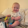baby, smiling, toys, pillow, bed, child, indoor, clothing, happy, face, person, infant, cute, sitting, playful, cushion, home, blanket, foot, light