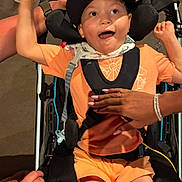 Lennox joined the competition — help win amazing prizes! child, wheelchair, orange_clothing, cap, smiling, happy, hands, support, indoor, person, seated, brace, disability, care, joyful, expression, shorts, bandana, black_cap, adult_hands