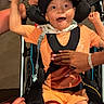 child, wheelchair, orange_clothing, cap, smiling, happy, hands, support, indoor, person, seated, brace, disability, care, joyful, expression, shorts, bandana, black_cap, adult_hands