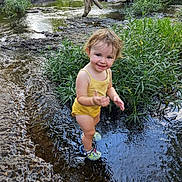 Piper joined the competition — help win amazing prizes! toddler, child, water, creek, nature, greenery, yellow_clothing, happy, smiling, outdoor, shoes, splashing, plants, logs, summer, adventure, exploring, young_child, river, playful