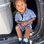 child, toddler, denim_clothing, curly_hair, sitting, car, car_interior, black_sneakers, socks, curious_expression, young_child, casual_wear, daylight, outdoor_light, car_door, small_person, candid, portrait, hands, footwear