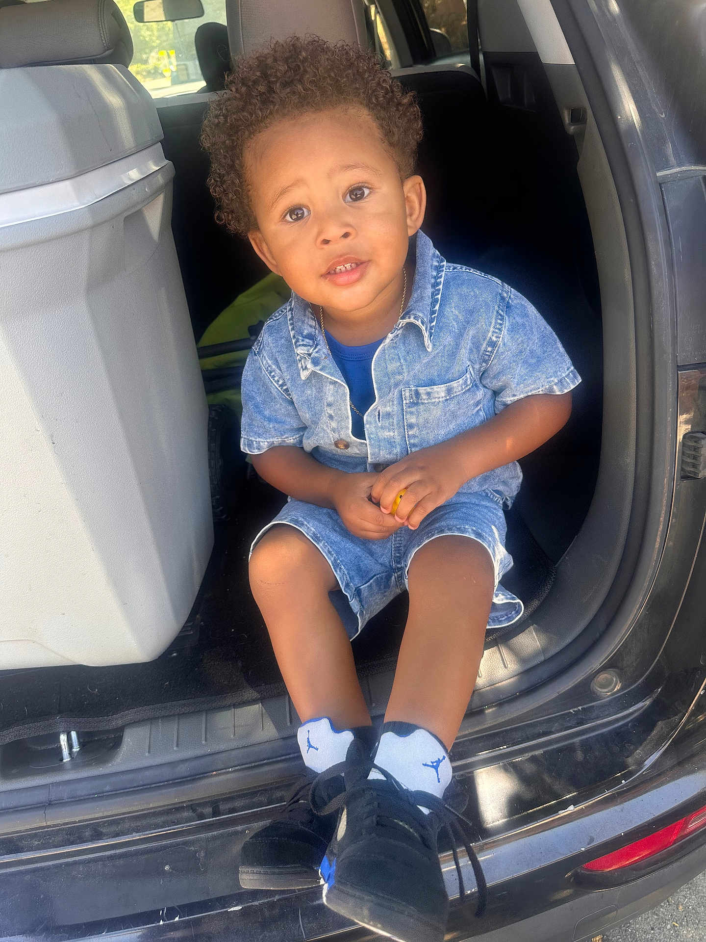 Christopher joined the competition — help win amazing prizes! child, toddler, denim_clothing, curly_hair, sitting, car, car_interior, black_sneakers, socks, curious_expression, young_child, casual_wear, daylight, outdoor_light, car_door, small_person, candid, portrait, hands, footwear