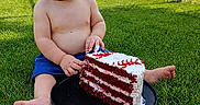 Walter is registered to the contest to win money with this photo: baby, child, grass, cake, red_velvet_cake, shirtless, blue_shorts, necklace, outdoor, daytime, fence, sports_field, food, plate, sitting, curious_expression, barefoot, summer, sky, greenery