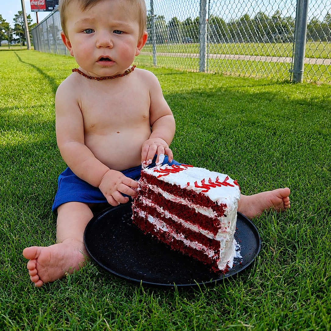 Walter is registered to the contest to win money with this photo: baby, child, grass, cake, red_velvet_cake, shirtless, blue_shorts, necklace, outdoor, daytime, fence, sports_field, food, plate, sitting, curious_expression, barefoot, summer, sky, greenery