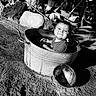 Theodore is registered to the contest to win money with this photo: toddler, child, metal_tub, water, outdoor, rocks, tree_trunk, garden_tools, shadow, sunlight, happy, smiling, playful, dirt, ground, bucket_lid, monochrome, black_and_white, cute, portrait