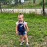 child, toddler, grass, outdoor, fence, denim_overalls, pink_shirt, shorts, sneakers, top_knot, trees, nature, person, summer, daylight, greenery, casual_clothing, standing, cute, young_child