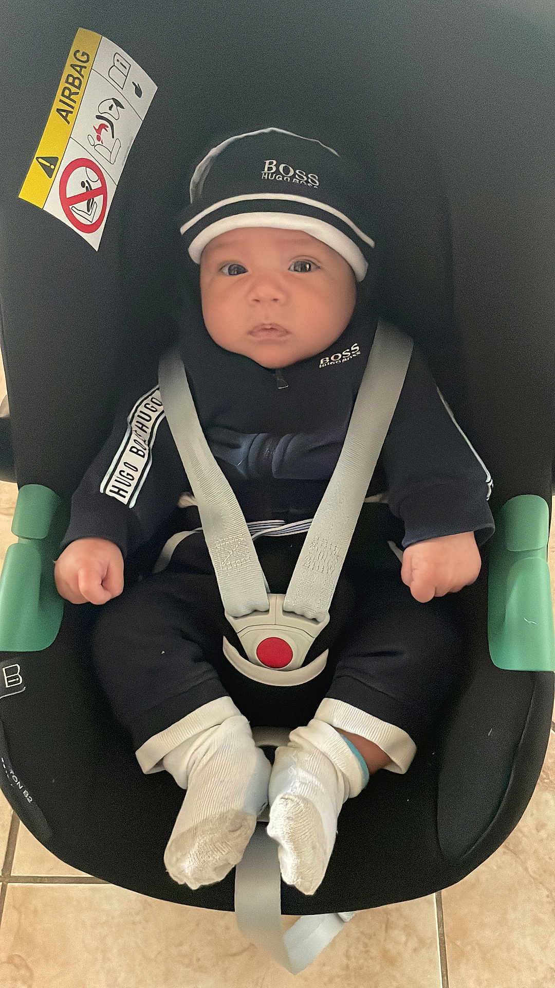 Jassim participe au concours pour gagner de l'argent avec cette photo : baby, car_seat, child, seat_belt, clothing, hat, socks, floor, indoor, person, face, hands, footwear, sitting, black_clothing, white_socks, tile_floor, safety, infant, hoodie