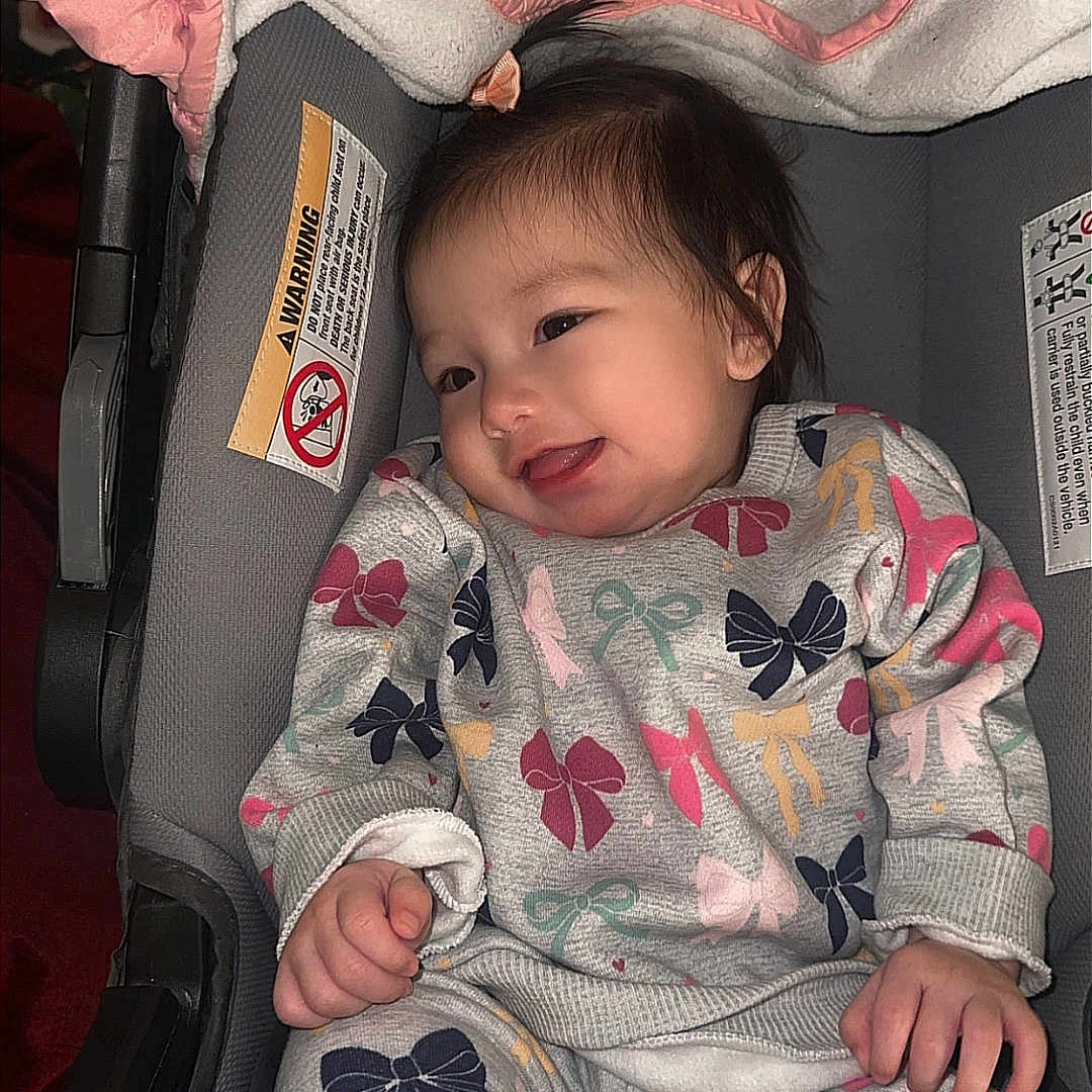 Kahmila is registered to the contest to win money with this photo: baby, car_seat, child, clothing, cozy, cute, face, gray, happy, indoor, infant, jacket, pattern, person, pink, relaxing, seat, smile, socks, young