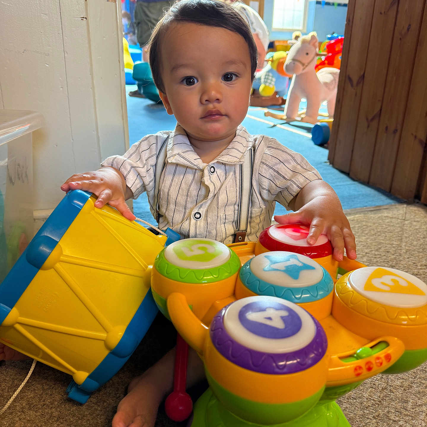 Freddie joined the competition — help win amazing prizes! carpet, child, colorful, cute, daylight, decorations, drum, foot, indoor, musical_toy, person, play_area, playroom, sitting, striped_shirt, suspenders, toddler, toy, window, wooden_door