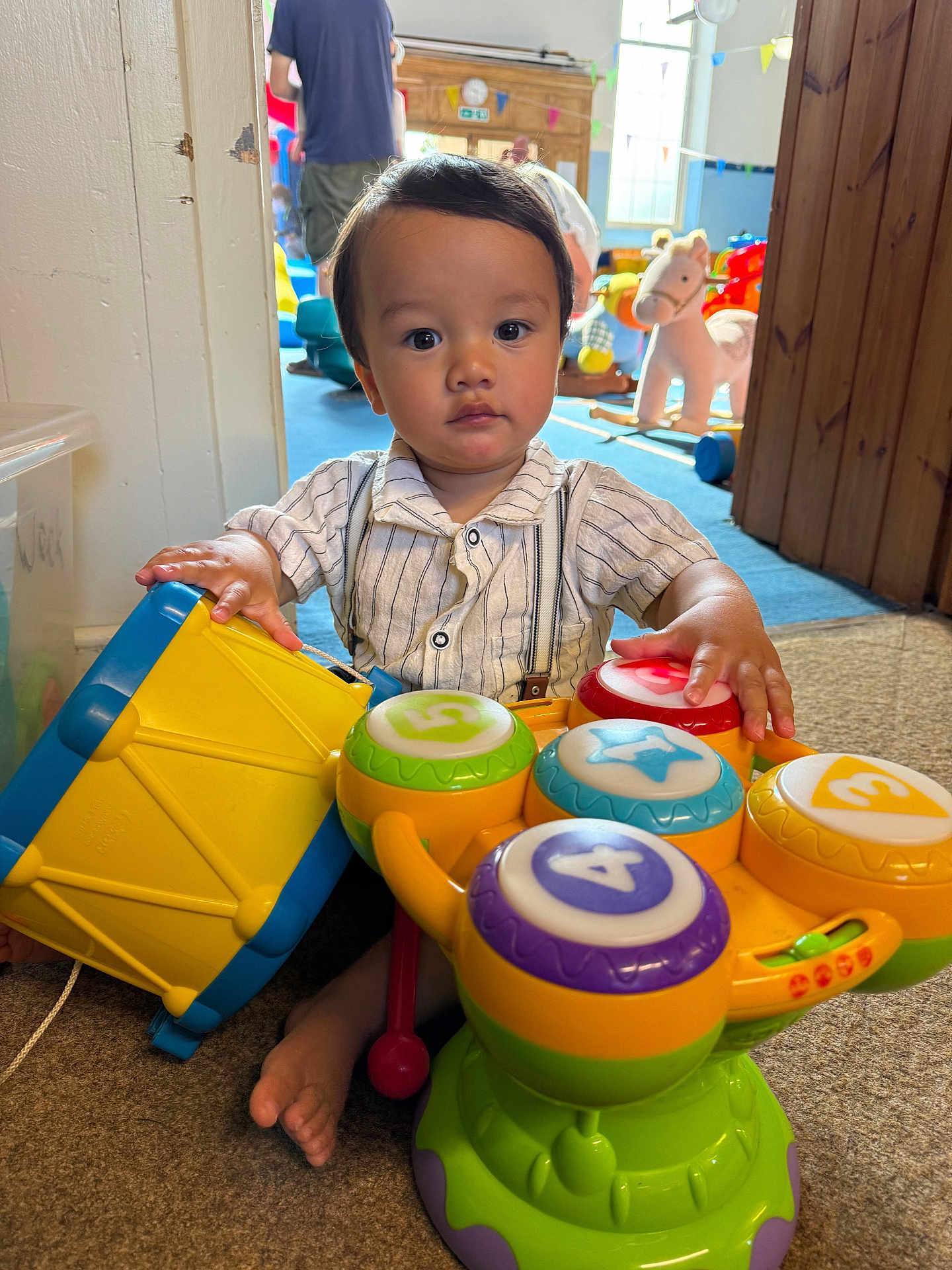 Freddie joined the competition — help win amazing prizes! carpet, child, colorful, cute, daylight, decorations, drum, foot, indoor, musical_toy, person, play_area, playroom, sitting, striped_shirt, suspenders, toddler, toy, window, wooden_door
